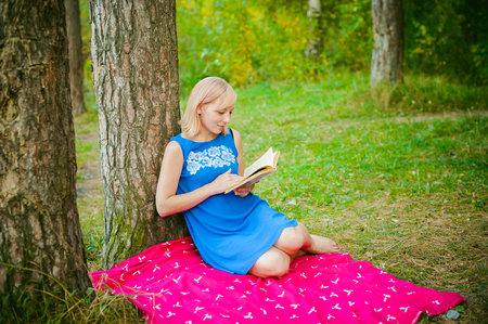 blonde girl in a blue dress sitting on a red blanket on top of the green grass, reading a book in the middle of pine forest treesの写真素材
