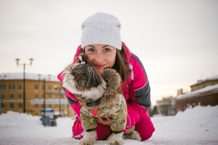 Walk in winter outdoors with dog breed Shih Tzu. A woman in bright red warm ski clothing walking in snow with your pet, little shih tzu dressed in overalls. care for animals loves playing with the dogの写真素材