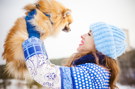 smiling woman holding her pet dog in his hands near face, overhead. Spitz breed dog playing with a woman walking outdoors winter day, warm clothing. love and care for the pet, dog walkingの写真素材
