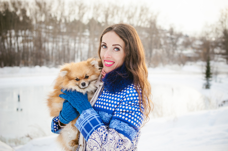 smiling woman holding her pet dog in his hands near face, hugging. Spitz breed dog playing with a woman walking outdoors winter day, warm clothing. love and care for the pet, dog walkingの写真素材