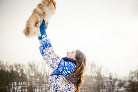 smiling woman holding her pet dog in his hands near face, tossing up overhead. Spitz breed dog playing with a woman walking outdoors winter day, warm clothing. love and care for the pet, dog walkingの写真素材