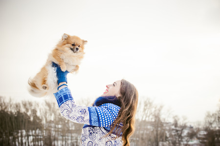 smiling woman holding her pet dog in his hands near face, tossing up overhead. Spitz breed dog playing with a woman walking outdoors winter day, warm clothing. love and care for the pet, dog walkingの写真素材