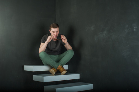studio photography young brutal guy. man in sunglasses, T-shirt, jeans and high boots on a background of black wall on the white steps. sitting on the steps, took off his glasses and holds them in his handの写真素材