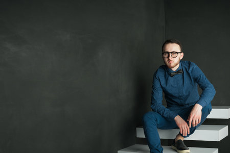 studio portrait of a young man on a dark wall background in a blue shirt and jeans. hipster man,...