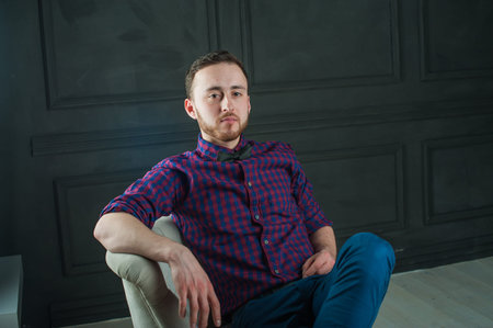 studio portrait of a young man on a dark wall background in a burgundy plaid shirt and blue jeans....