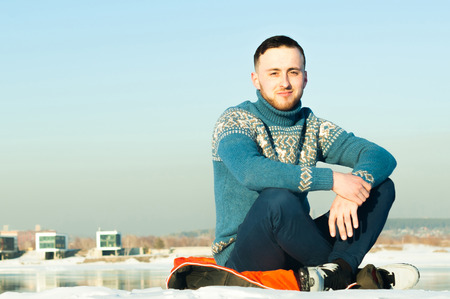 man in warm sweater with a beard is walking on the street in the winter in a warm sunny day at the river against the backdrop of the city, sitting in the snow on the beach with a view of the opposite shoreの写真素材