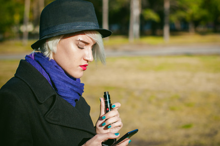 portrait of young beautiful woman with white hair, in a black coat, a skirt and a black hat, smoking an electronic cigarette, blowing the smoke vaporの写真素材