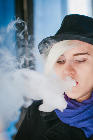 portrait of young beautiful woman with white hair, in a black coat, a skirt and a black hat, smoking an electronic cigarette, blowing the smoke vaporの写真素材