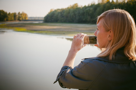 pretty blonde girl in sneakers, nylon stockings and coat. walks and photographed on a smartphone selfi on the stone shore of the lake in the last sunshine of the departing summer.の写真素材