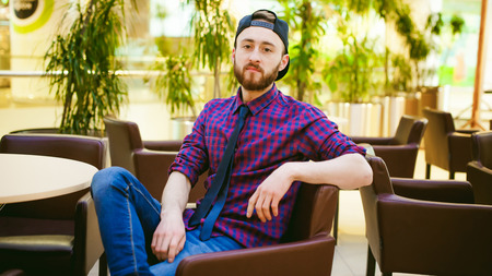 Portrait of a young man with a beard, dressed in jeans and a plaid shirt, tie and cap, sitting on a chair at the coffee shop in the mallの写真素材