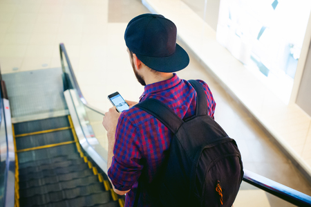 young man with a beard in a plaid shirt and a tie and cap, riding on an escalator in the mall, looking at smartphoneの写真素材
