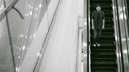 young man with a beard in a plaid shirt and a tie and cap, riding on an escalator in the mallの写真素材