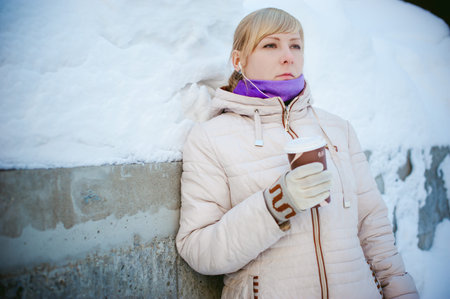 young woman dressed in warm clothes drinking takeaway coffee from the cup on the street in winter