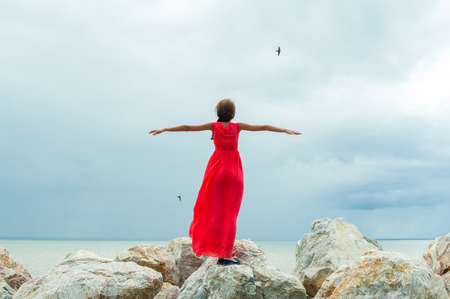 girl in red dress standing on the rocks, his arms outstretched like wings of a bird in flight, against the seaの写真素材