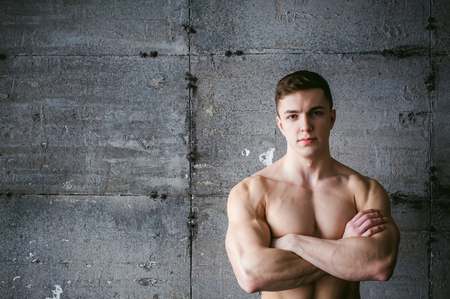 Young sexy male bodybuilder athlete with a bare torso against a gray brutal concrete wall, portrait of a studio of a light-skinned strong man crossed powerful arms on his chestの写真素材