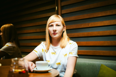 Young blonde woman wearing white T-shirt with print, girl Drinks from a white porcelain cup holding it in his hands, sitting at a table in a cafe, background stylized Wood texture lit up with warm lightの写真素材