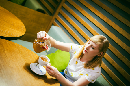 Young blonde woman wearing white T-shirt with print, girl pours a fruit drink from a glass jug, into a white cup, sitting at a table in a cafe, background stylized Wood texture lit up with warm lightの写真素材