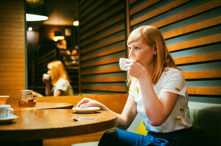Young blonde woman wearing white T-shirt with print, visited cafe lunch time, drinking fruity hot drink from glass teapot white cup hand holding, background stylized tree table lit up with warm lightの写真素材
