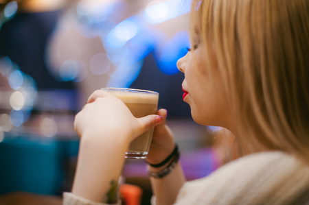 Cute young blonde woman drinks hot coffee from a glass cup in the interior of a coffee house. Abstract blur, out of focusの写真素材