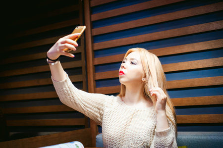 young blond woman in a knitted sweater, making a selfie photo on a smartphone, sitting at a table in a cafe, spending time alone, doing internet communicationの写真素材