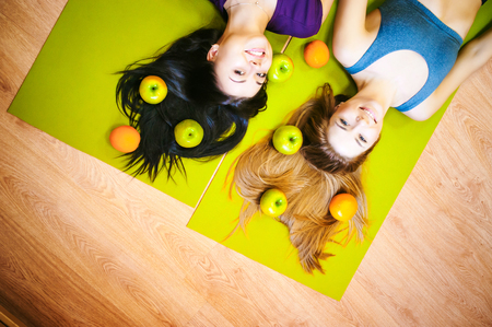 Two young athletic physique women in gym Lie on the floor on yoga mats. Fitness girl with fruits, lifestyle in losing weight and diets, proper nutrition, exerciseの写真素材
