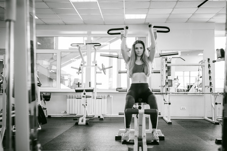 girl in a bright white gym. A blonde woman doing a strength exercise for a group of muscles on the hands of a simulator. Healthy way of life, good spiritsの写真素材