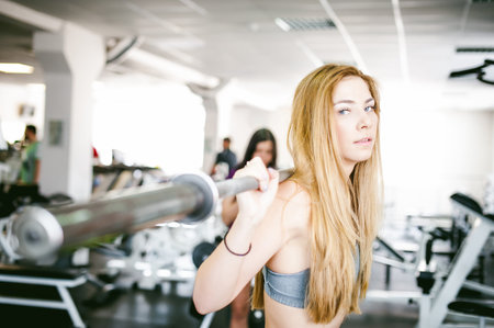 girl in bright white gym. Athletic slender sexy blonde woman doing strength exercise for group of leg muscles and buttocks squatting with lightweight barbell barb. Healthy way of life, good spiritsの写真素材