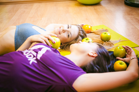 Two young athletic physique women in gym Lie on the floor on yoga mats. Fitness girl with fruits, lifestyle in losing weight and diets, proper nutrition, exerciseの写真素材