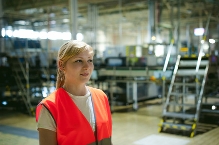 Portrait of a female employee in an orange robe vest in the working space of a production facility, supervises and controls the line in food production, against the background of equipmentの写真素材