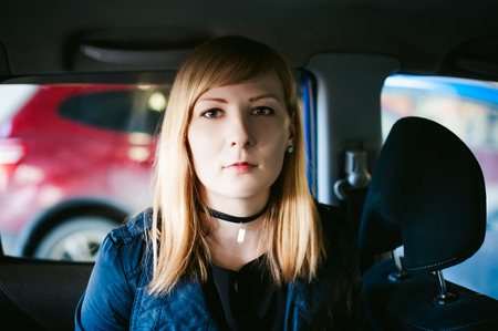 Portrait of a blond woman sitting in a car in the back seat, dressed in a raincoat and a checker on the neck, the picture is taken through the back of the carの写真素材