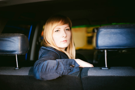 Portrait of a blonde woman sitting in a car in the back seat, placing elbow on the back of the seat, picture taken through the back of the carの写真素材