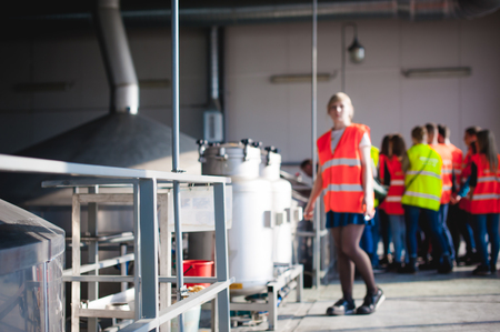 blonde woman in overalls, on the tour of the brewery plant in industrial premises, amid crowds of people in vests, inspecting a factory for training purposesの写真素材