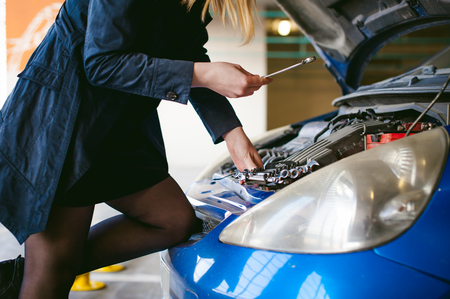 woman near car's hood. young blonde in covered parking of shopping center, stands near car with engine compartment bonnet raised, solves problem with engine malfunction, using carabiner wrench toolの写真素材