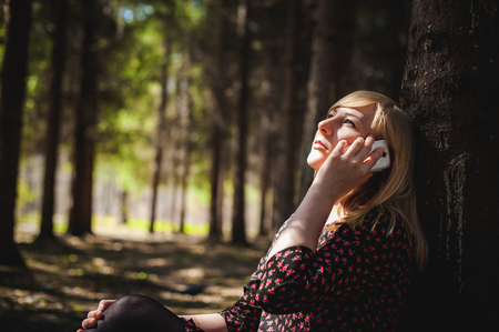 Walk girl coniferous forest park. blonde woman dressed summer dress, black pantyhose and sneakers with white sole, resting nature enjoying warm sunny day. Sits under tree alone, Speaks cell phoneの写真素材