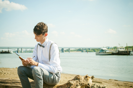 young skinny man, elegantly stylishly dressed in white shirt, gray trousers with suspenders and bow tie. young guy on pier on background of river and boats, takes notes of pen on sheet in notebookの写真素材