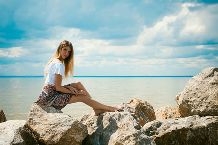 Portrait of slender young woman on stones near the sea. Beautiful girl is resting on coast, enjoying outdoor recreationの写真素材