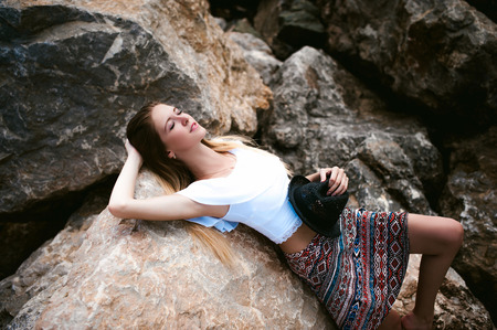 Portrait of slender young woman on stones near sea. Beautiful girl rests on coast, enjoying outdoor recreation, holds a black hat in her handsの写真素材
