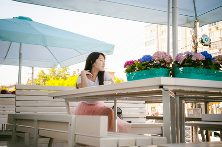 Cute asian young woman in summer cafe outdoors. girl In white T-shirt, with long hair in simple light cozy interior of restaurant Urban style, drinking coffee drink, sitting at table with flowersの写真素材