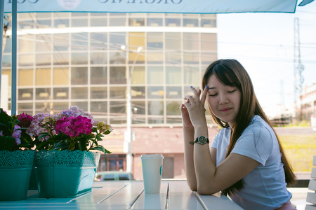 Cute asian young woman in summer cafe outdoors. girl In white T-shirt, with long hair in simple light cozy interior of restaurant Urban style, drinking coffee drink, sitting at table with flowersの写真素材