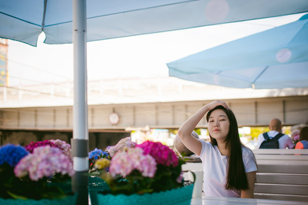 Cute asian young woman in summer cafe outdoors. girl In white T-shirt, with long hair in simple light cozy interior of restaurant Urban style, drinking coffee drink, sitting at table with flowersの写真素材
