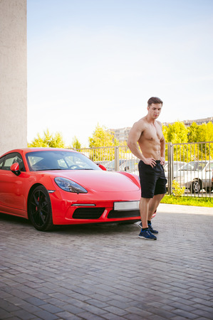 Handsome sexy male athlete man with naked torso in yard of house in parking lot Near an expensive prestigious red sports racing car, an early sunny summer morningの写真素材
