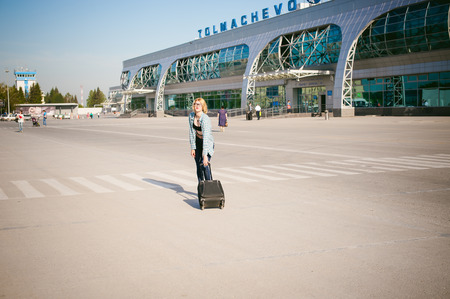 young slim woman dressed in blue checked shirt, jeans, Glasses. Will depart at dawn on summer day on freedom trip with black travel suitcase Against the airport buildingのeditorial素材