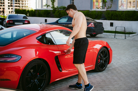 Handsome sexy male athlete man with naked torso in yard of house in parking lot Near an expensive prestigious red sports racing car, an early sunny summer morningの写真素材