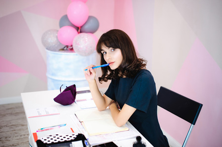 Portrait of a beautiful business woman making notes in a notebook. Holds a pen, sitting in the workplace in a pink officeの写真素材