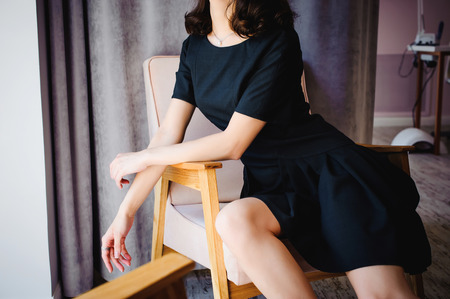 Young attractive woman with long legs in black elegant dress, sits in chair near window in interior of room.の写真素材