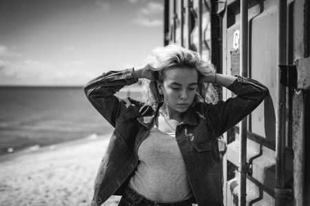 young beautiful woman in jeans clothes outdoors. portrait of a girl with freckles on her face, stylish girl on a freight containers background, on a sunny summer day. wind develops her hairの写真素材