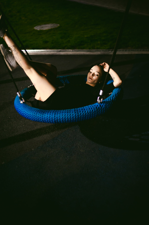 young woman with beautiful feet dressed in white sneakers and black dress lies in a swing in the yard, against a backdrop of urban landscapes. Street fashion. night dark styleの写真素材