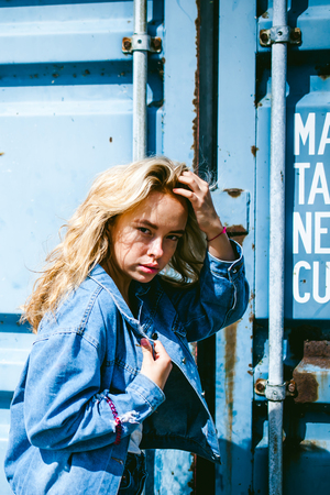 young beautiful woman in jeans clothes outdoors. portrait of a girl with freckles on her face, stylish girl on a freight containers background, on a sunny summer day. wind develops her hairの写真素材