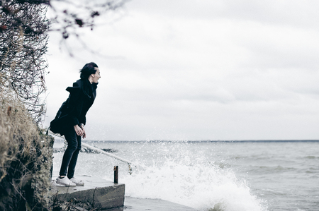 fashionable portrait of a young brunette woman in black clothes, jeans T-shirt, coat and sunglasses, in a Gothic style sad mood. on an empty beach, on an autumn cold overcast day. waves are ragingの写真素材
