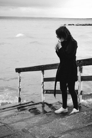 fashionable portrait of a young brunette woman in black clothes, jeans T-shirt, coat and sunglasses, in a Gothic style sad mood. on an empty beach, on an autumn cold overcast dayの写真素材
